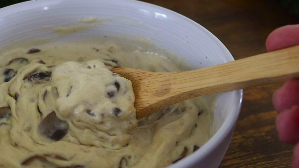 A bowl filled with creamy mushroom sauce, with visible mushroom pieces. A wooden spoon is stirring the mixture inside the white bowl, which is set on a wooden surface.