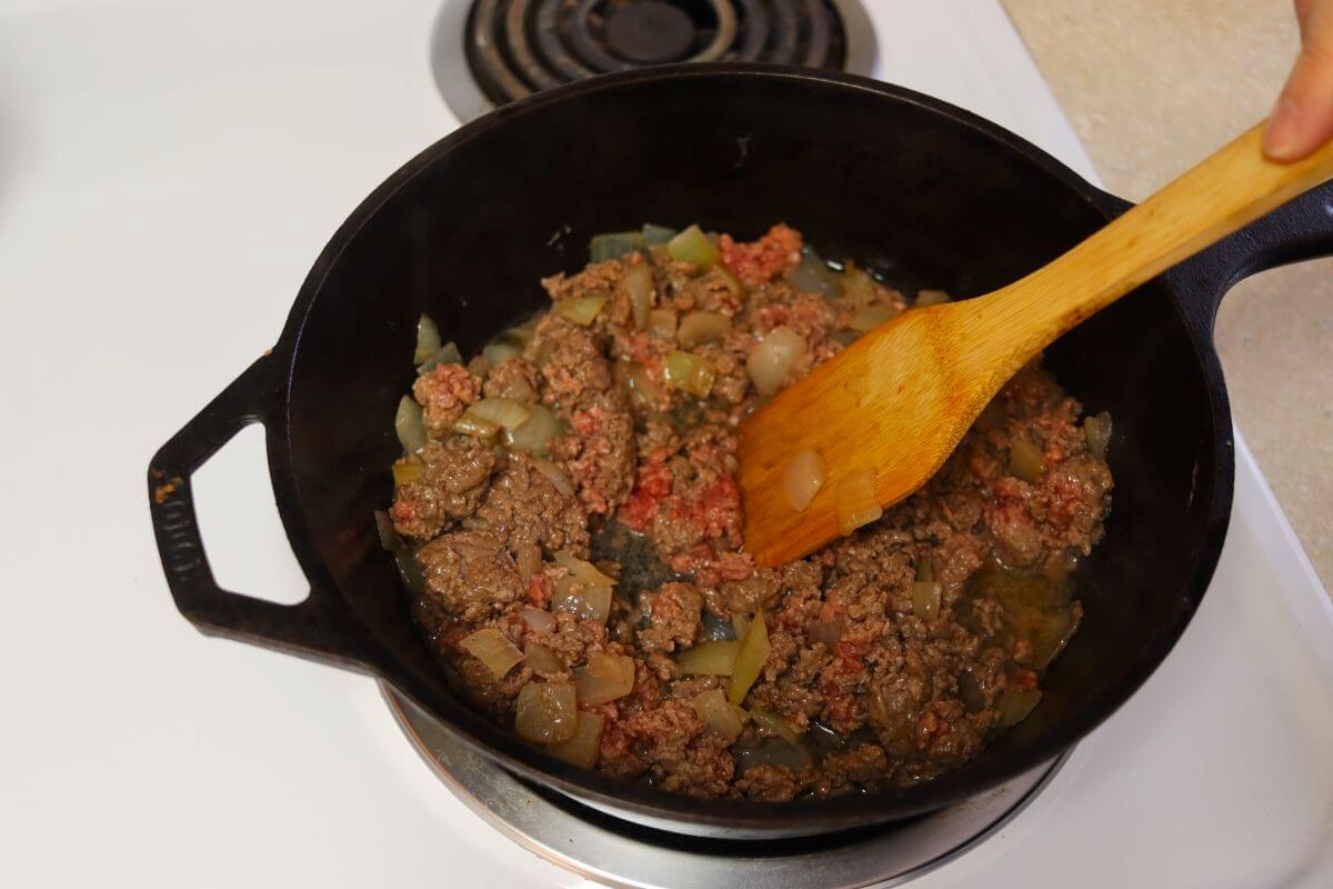 Partaily cooked ground beef with onions in it cooking in a cast iron skillet with a wooden spoon sitting in it.