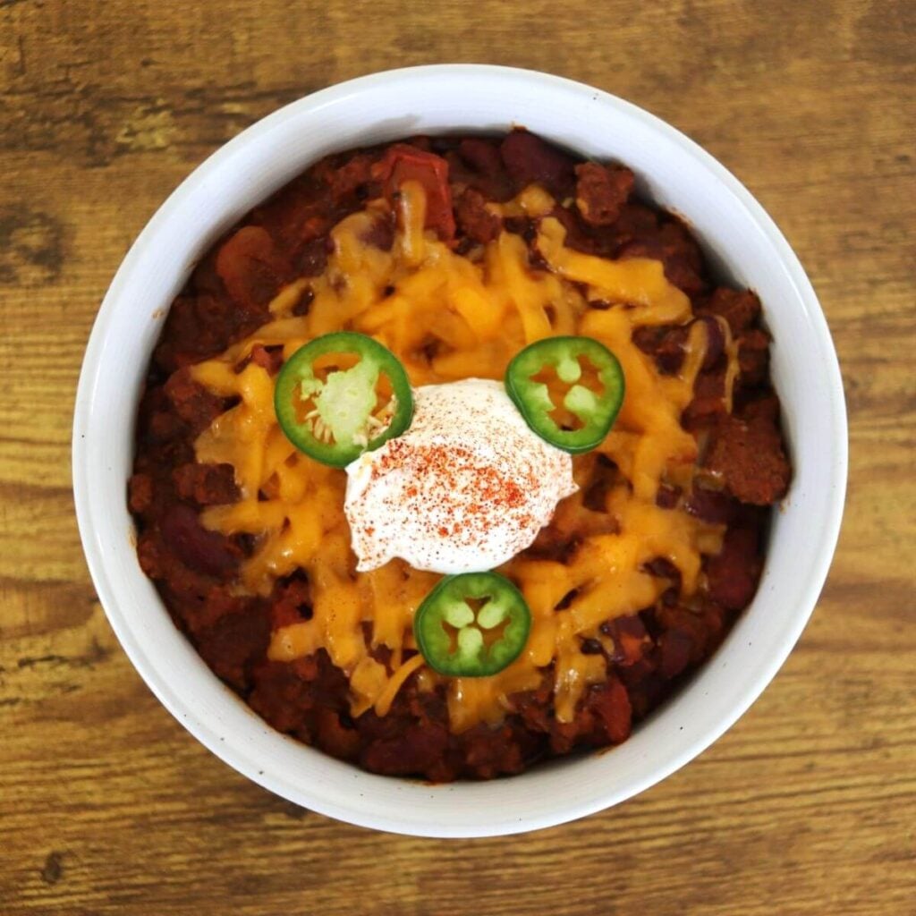 White bowl full of chili topped with melted yellow cheese, a scoop of sour cream, sprinkled with paprika, and 3 slices of fresh jalapeno.