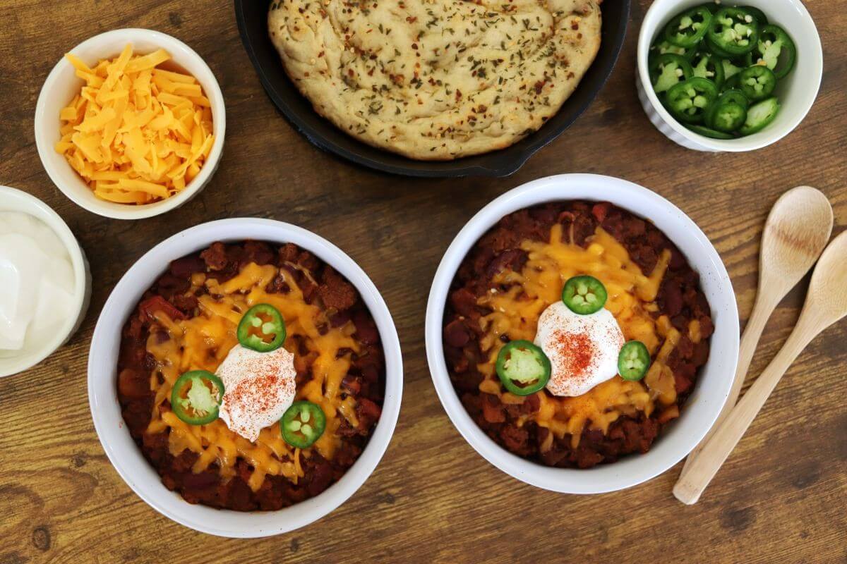 Two bowls of chili topped with shredded cheese, sour cream, sliced jalapeños, and paprika. Surrounding them are wooden spoons, a bowl of shredded cheese, sour cream, sliced jalapeños, and a round herb flatbread on a wooden table.
