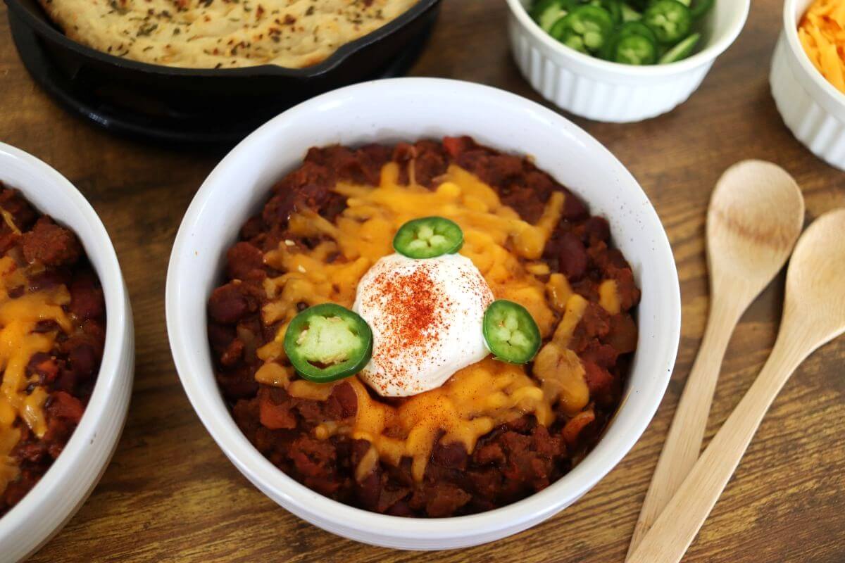 A bowl of chili topped with shredded cheddar cheese, a dollop of sour cream, three jalapeño slices, and a sprinkle of paprika. A dish of sliced jalapeños, cornbread, spoons, and shredded cheese are nearby.