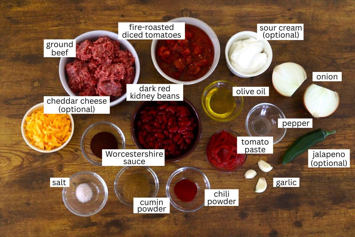 A variety of chili ingredients on a wooden surface, each labeled: ground beef, dark red kidney beans, fire-roasted diced tomatoes, tomato paste, Worcestershire sauce, olive oil, garlic, onion, pepper, salt, cumin powder, chili powder, jalapeno (optional), cheddar cheese (optional), and sour cream (optional).