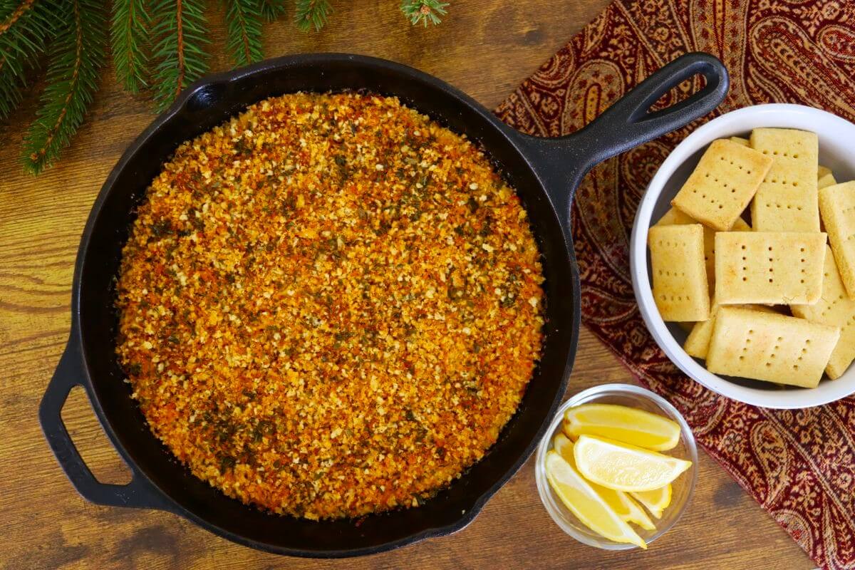 A cast iron pan filled with a crumbly, golden-orange dish sits on a wooden table next to a bowl of rectangular shortbread cookies and a small glass bowl containing lemon wedges.