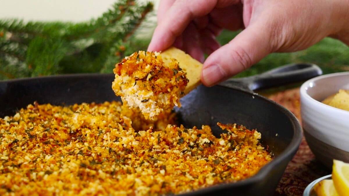 A hand dips a homemade cracker into a skillet filled with a golden, crispy breadcrumb-topped dish, with fresh herbs and lemon wedges visible in the background.