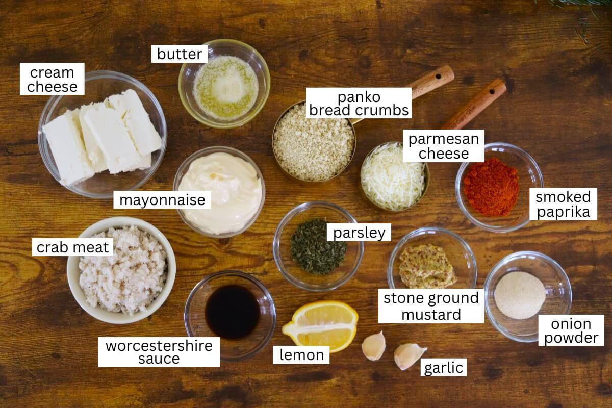 Labeled ingredients in small bowls for a crab cake recipe, including cream cheese, butter, panko bread crumbs, parmesan, smoked paprika, mayonnaise, crab meat, parsley, mustard, lemon, Worcestershire, onion powder, and garlic.