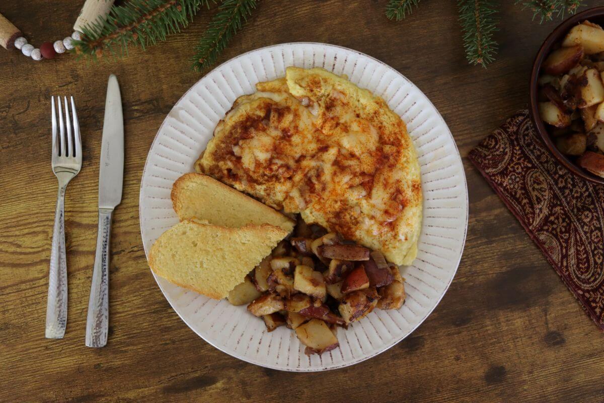 A plate with a cheesy omelette, two slices of toast, and a serving of diced, cooked potatoes. Silverware is placed to the left, and evergreen sprigs decorate the wooden table.