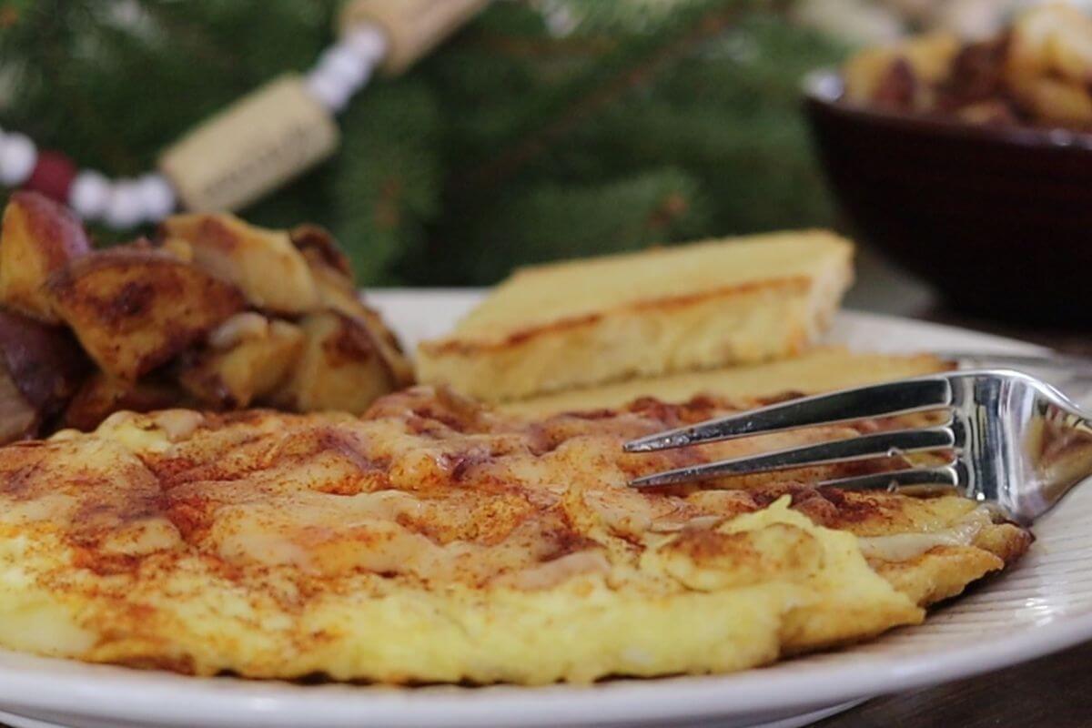 A close-up of a plate with a fluffy omelette, a serving of roasted potatoes, and a slice of semolina toast. A fork rests on the omelette. A blurred, festive background is visible.