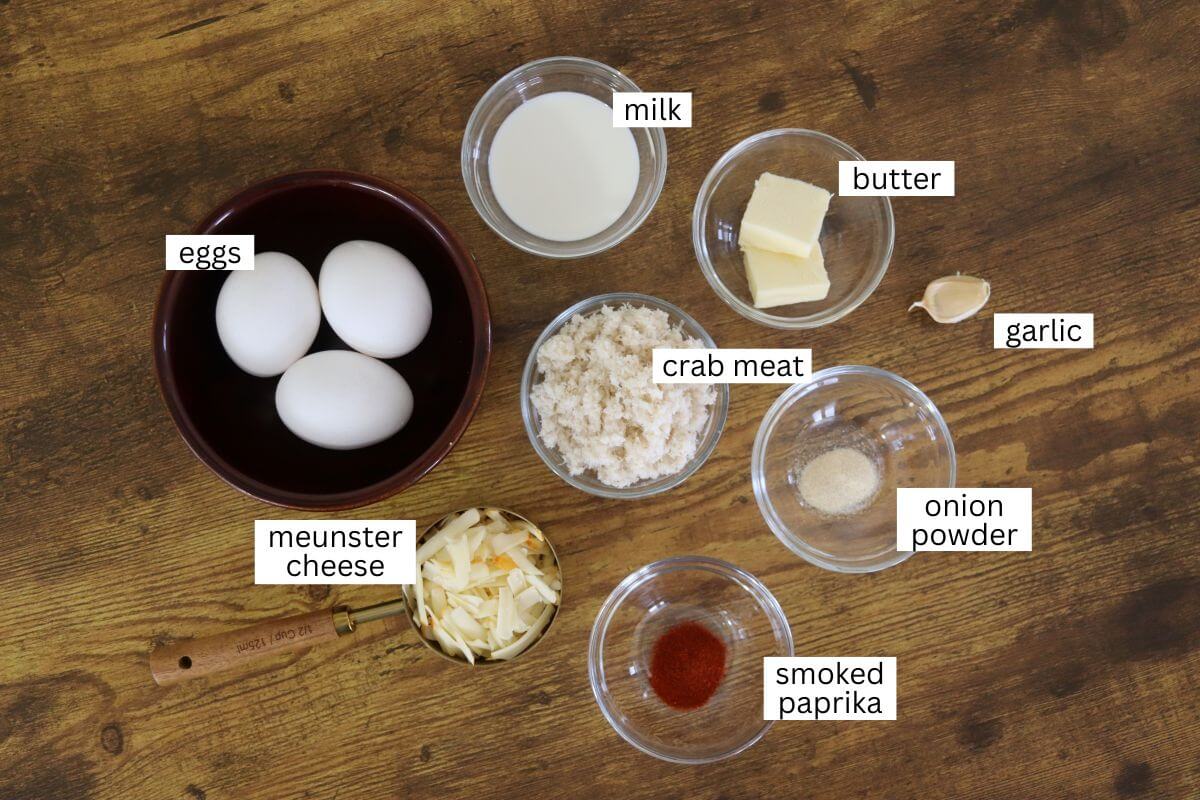 A wooden table with labeled ingredients in bowls: eggs, milk, butter, garlic, crab meat, onion powder, smoked paprika, and shredded muenster cheese.