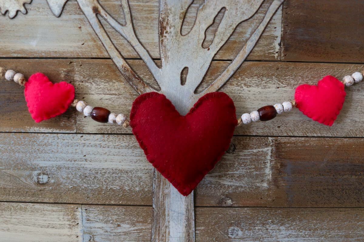 A garland of red and pink fabric hearts with beads is hung against a rustic wooden background with a carved tree design.