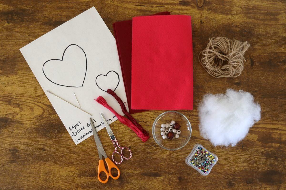 Craft materials on a wooden table: two sheets of paper with heart outlines, scissors, red thread, red felt, twine, stuffing, wooden beads in a bowl, and a container of colorful pins.