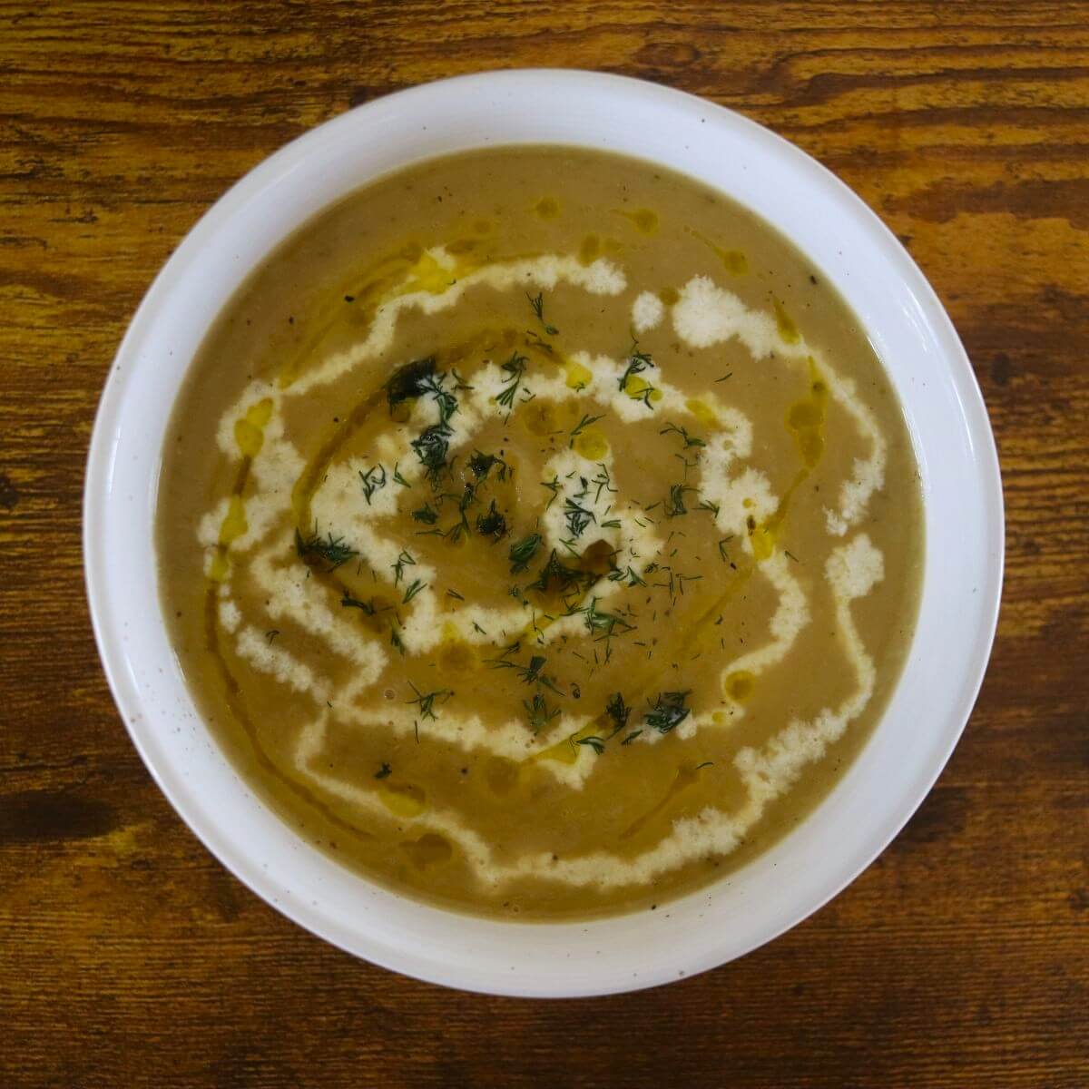 Creamy, light green soup in a white bowl with swirls of cream and olive oil and chopped fresh dill on top.