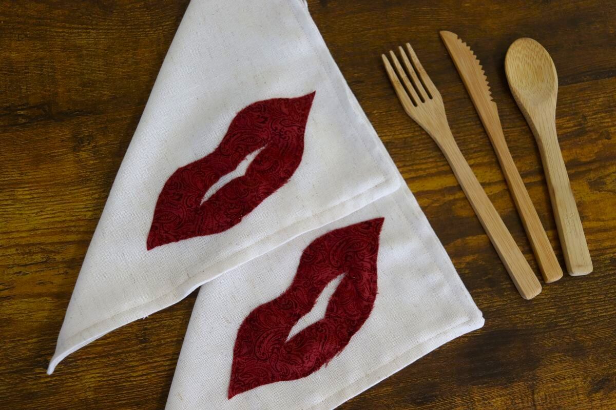 Two white cloth napkins with red lips designs are folded next to wooden cutlery, including a fork, knife, and spoon, on a wooden surface.