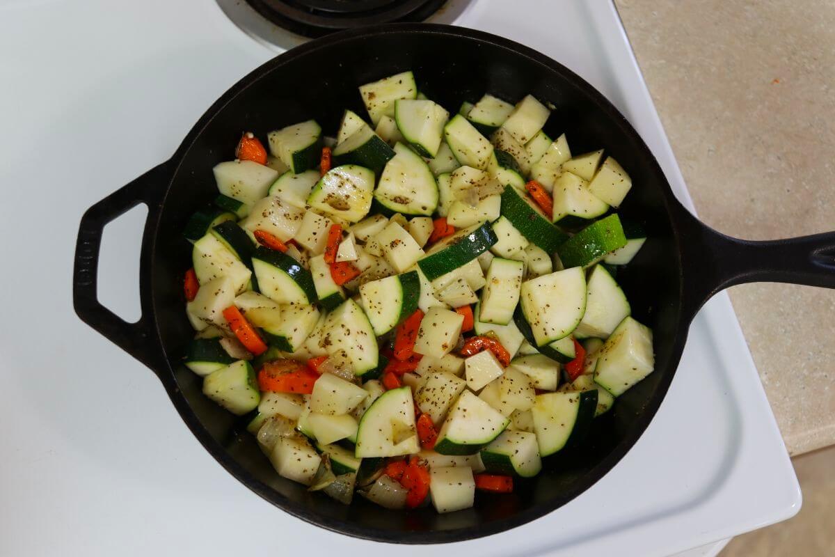 Chopped zucchini, potatoes, carrots, and onions with seasoning on them in a cast iron pot.