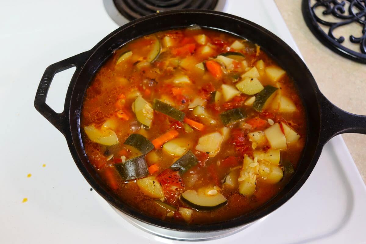 Vegetable soup in a cast iron pot that includes zucchini, carrots, onions, potatoes, and tomatoes.