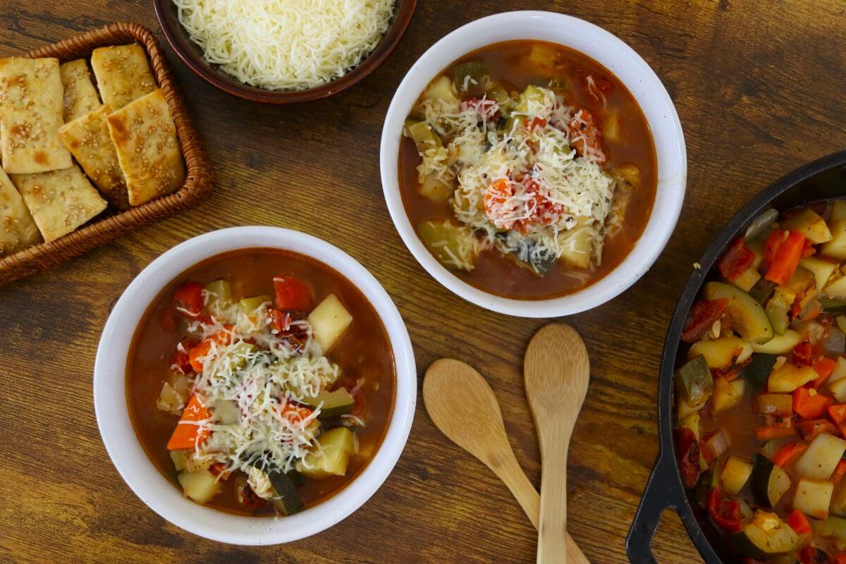 Two bowls of vegetable soup topped with shredded cheese sit on a wooden table, with wooden spoons beside them. Nearby are a basket of crackers, a bowl of extra cheese, and a pan of more soup.
