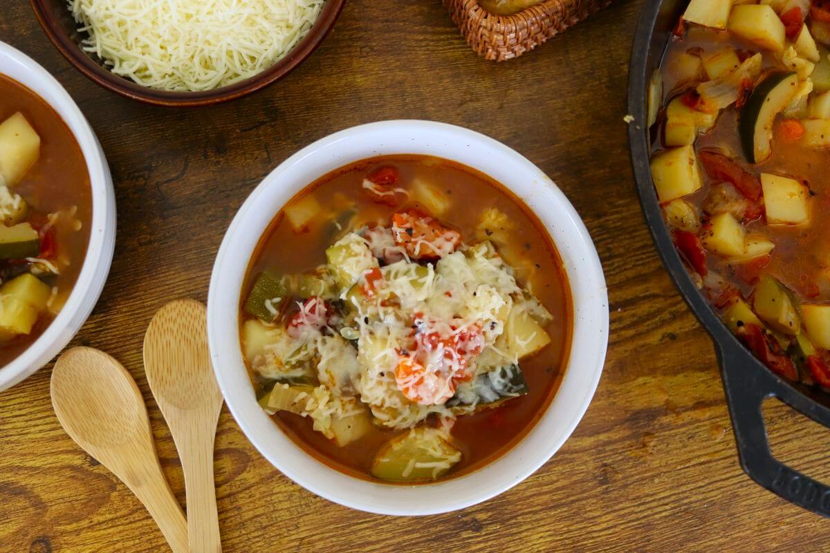 A bowl of vegetable soup topped with shredded cheese sits on a wooden table next to two wooden spoons, a pot of soup, and a bowl of grated cheese.
