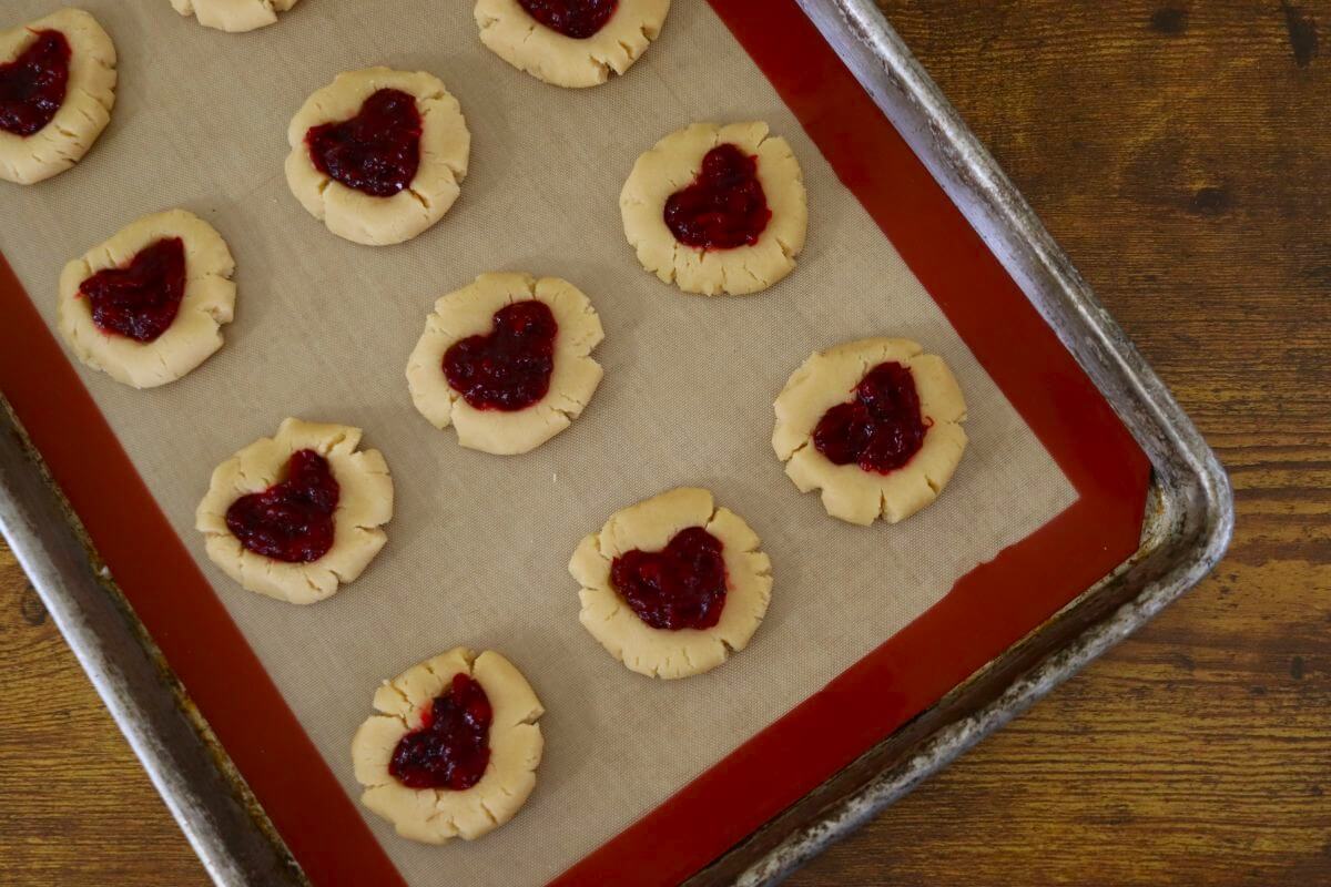 Flat, round cookies with jam filled heart shapes on them on a silicone lined baking sheet.