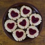 Seven round cookies with jam filled heart shapes on top on a plate.
