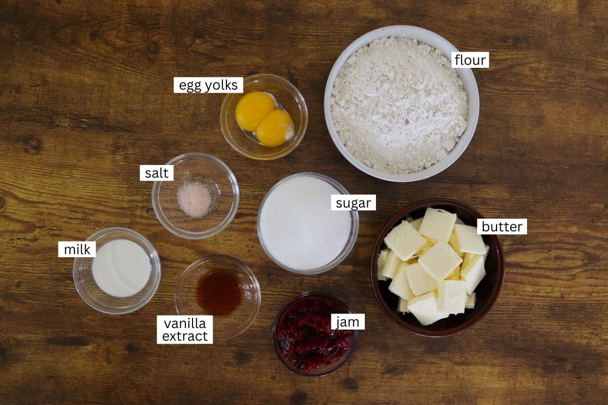 A wooden table with labeled ingredients in bowls: flour, butter cubes, sugar, jam, milk, vanilla extract, egg yolks, and salt, all arranged for baking.