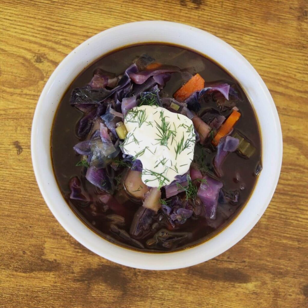 A bowl of soup that has red cabbage, carrots, and potatoes in it, topped with a dollop of sour cream and chopped fresh dill.