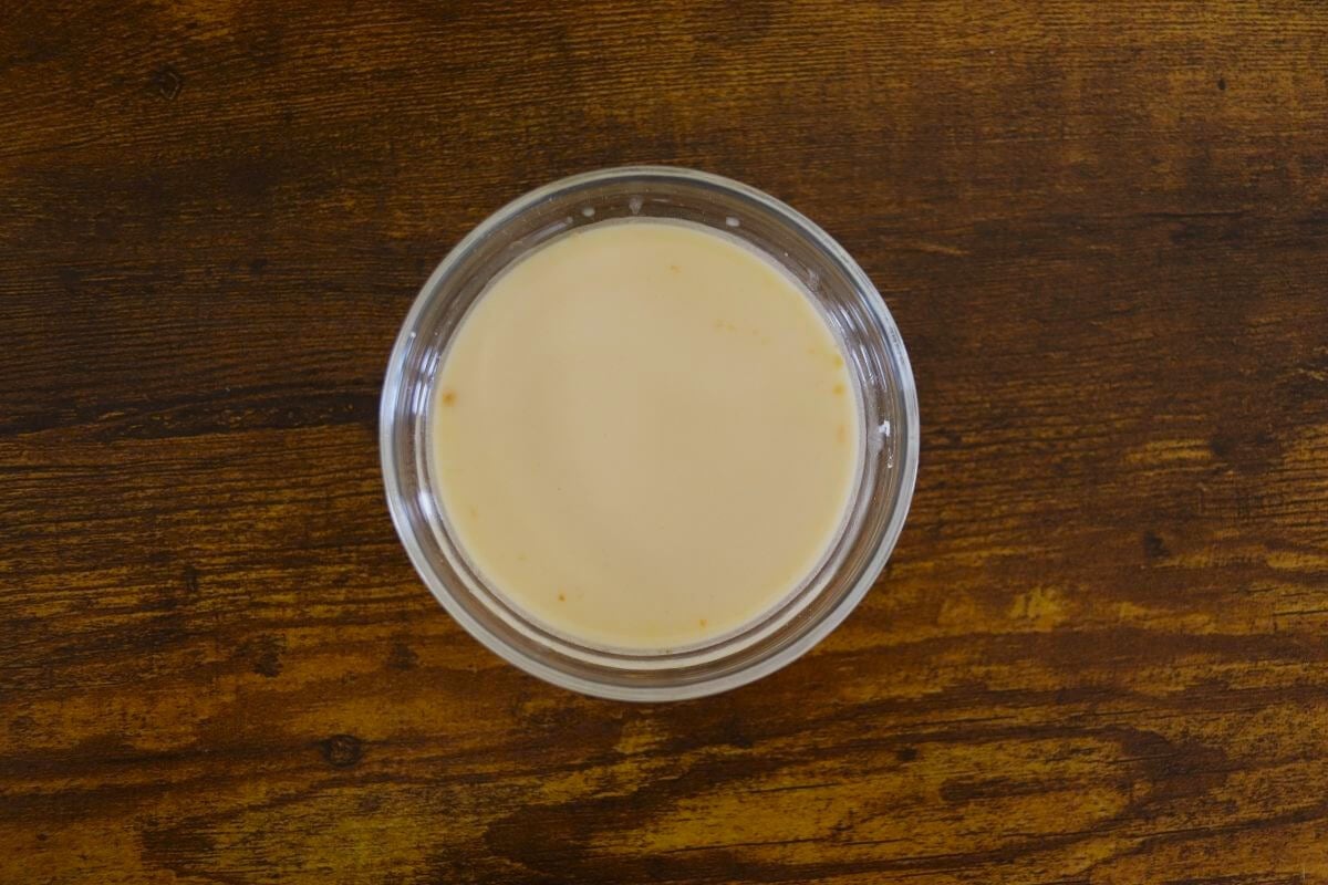 A small glass dish of a yellow tinted creamy liquid on a wooden surface.