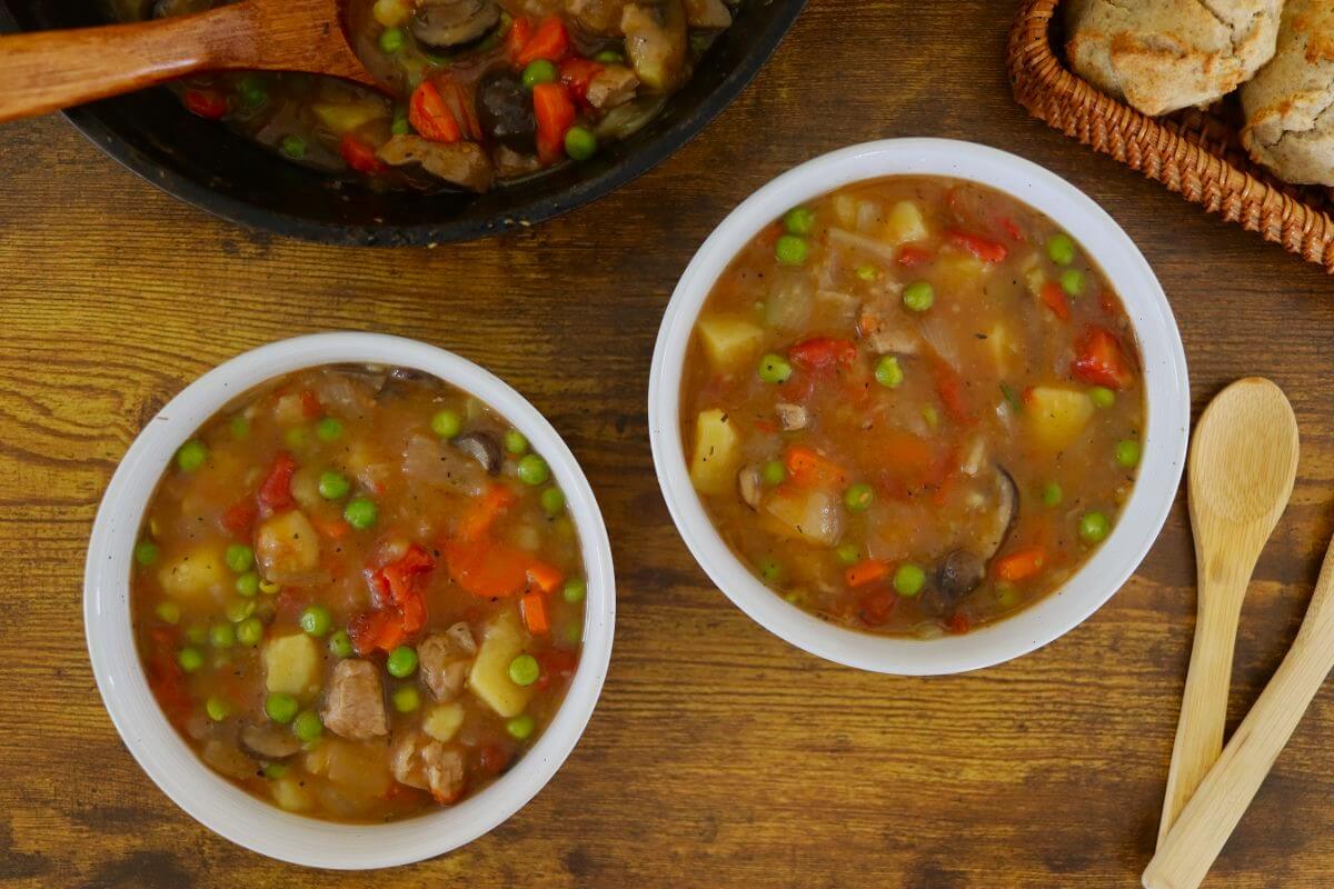 Two bowls of chunky pork stew with carrots, peas, potatoes, and mushrooms sit on a wooden table next to wooden spoons, a skillet with more stew, and a basket of biscuits.