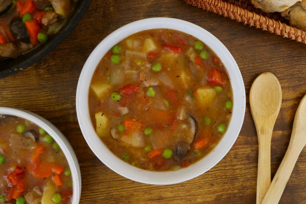 A bowl of pork stew with peas, carrots, potatoes, and mushrooms sits on a wooden table beside wooden spoons and a basket. Another partial bowl of stew is visible in the corner.