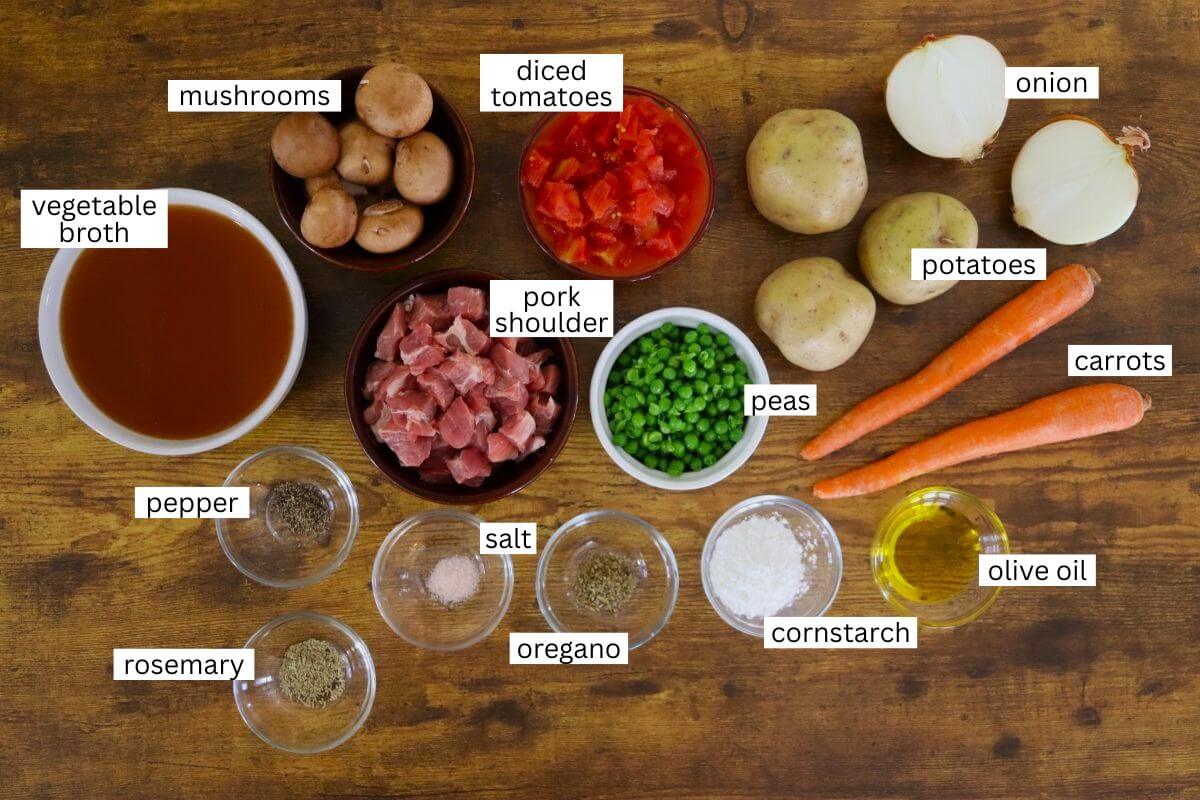 A variety of labeled stew ingredients including mushrooms, diced tomatoes, onion, potatoes, carrots, pork shoulder, peas, vegetable broth, pepper, rosemary, salt, oregano, cornstarch, and olive oil on a wooden surface.