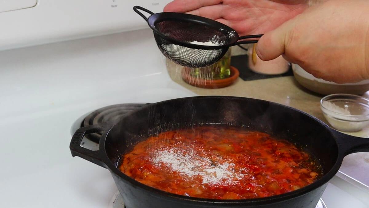 A hand tapping flour from a sieve style strainer into a cast iron skillet full of red broth.