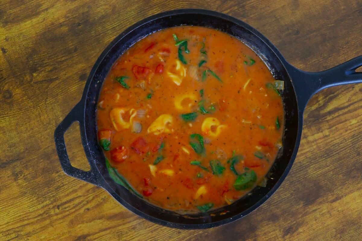 Cast iron skillet full of tomato based soup with spinach, onions, and tortellini visible in it sitting on a wooden surface.