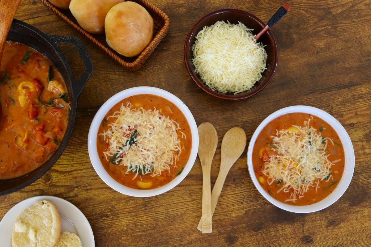 Two bowls of rustic tortellini soup topped with shredded cheese sit on a wooden table, alongside a pot of soup, a bowl of extra cheese, two wooden spoons, bread rolls, and split bread on a plate.