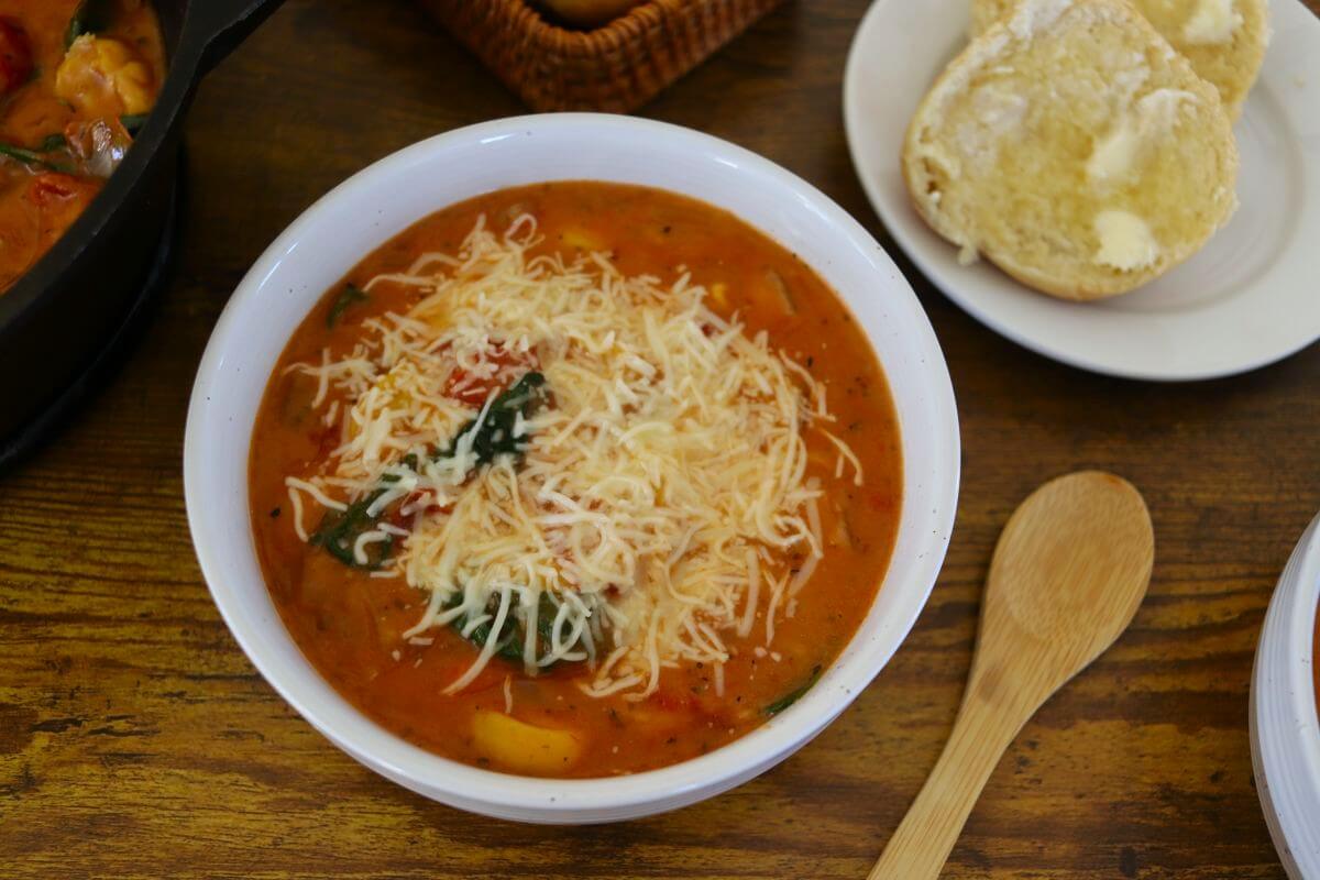A bowl of tomato-based soup topped with shredded cheese and greens, placed on a wooden table next to a buttered bread roll, a wooden spoon, and a woven basket.