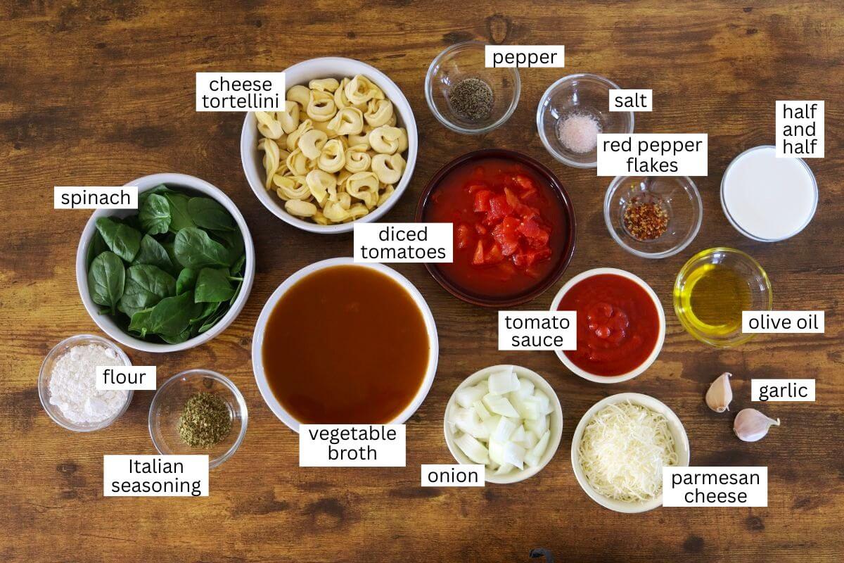 A wooden table with labeled bowls of ingredients: cheese tortellini, spinach, flour, Italian seasoning, vegetable broth, diced tomatoes, tomato sauce, onion, parmesan cheese, garlic, olive oil, red pepper flakes, salt, pepper, half and half.