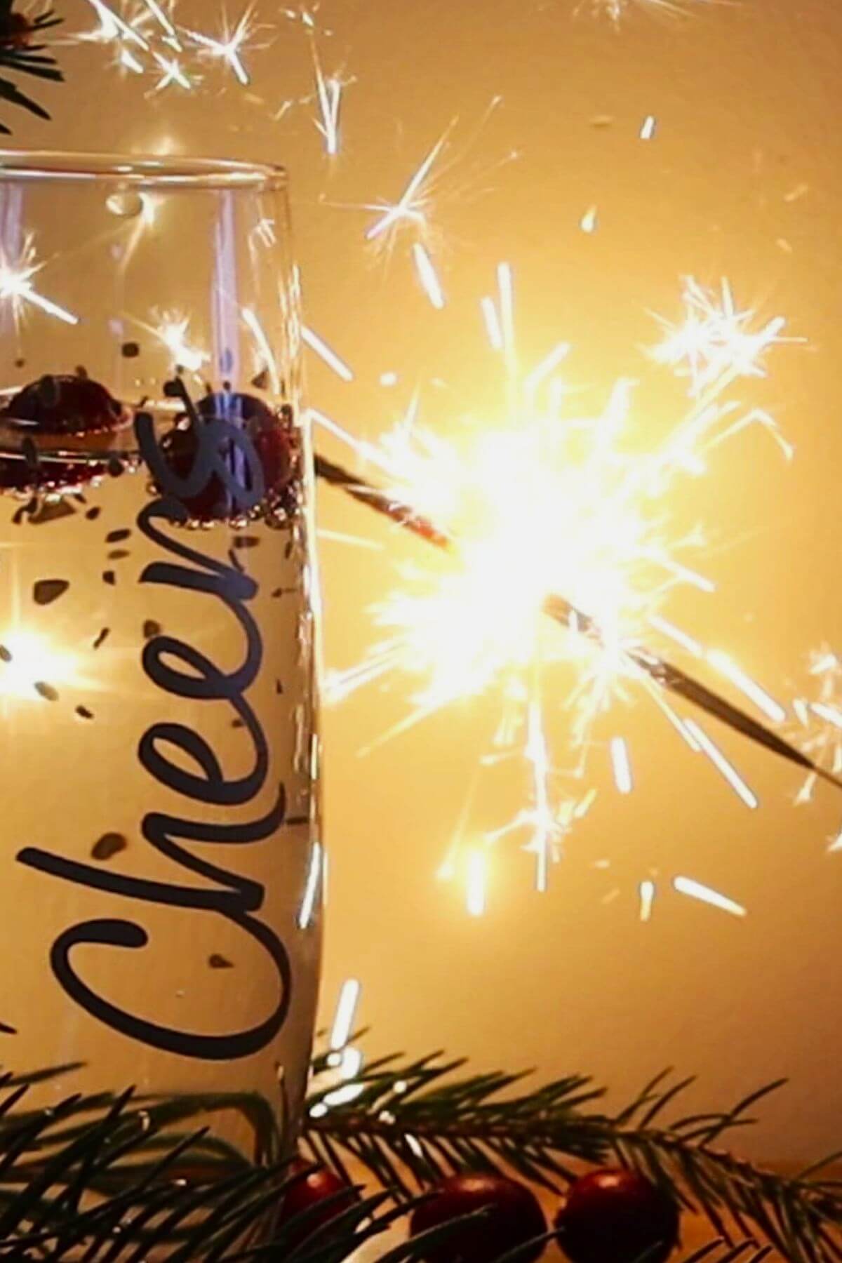 A glass of champagne with the word Cheers on it is in focus, adorned with red berries. A lit sparkler glows brightly in the background, creating a festive atmosphere. Pine branches decorate the bottom of the image.