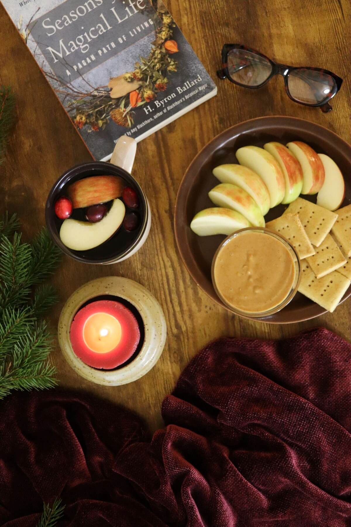 A cozy scene with apple slices and crackers beside a bowl of peanut butter, a mug of mulled drink with fruit, a lit candle, a book, reading glasses, and a red blanket on a wooden table. Pine branches are in the corner.