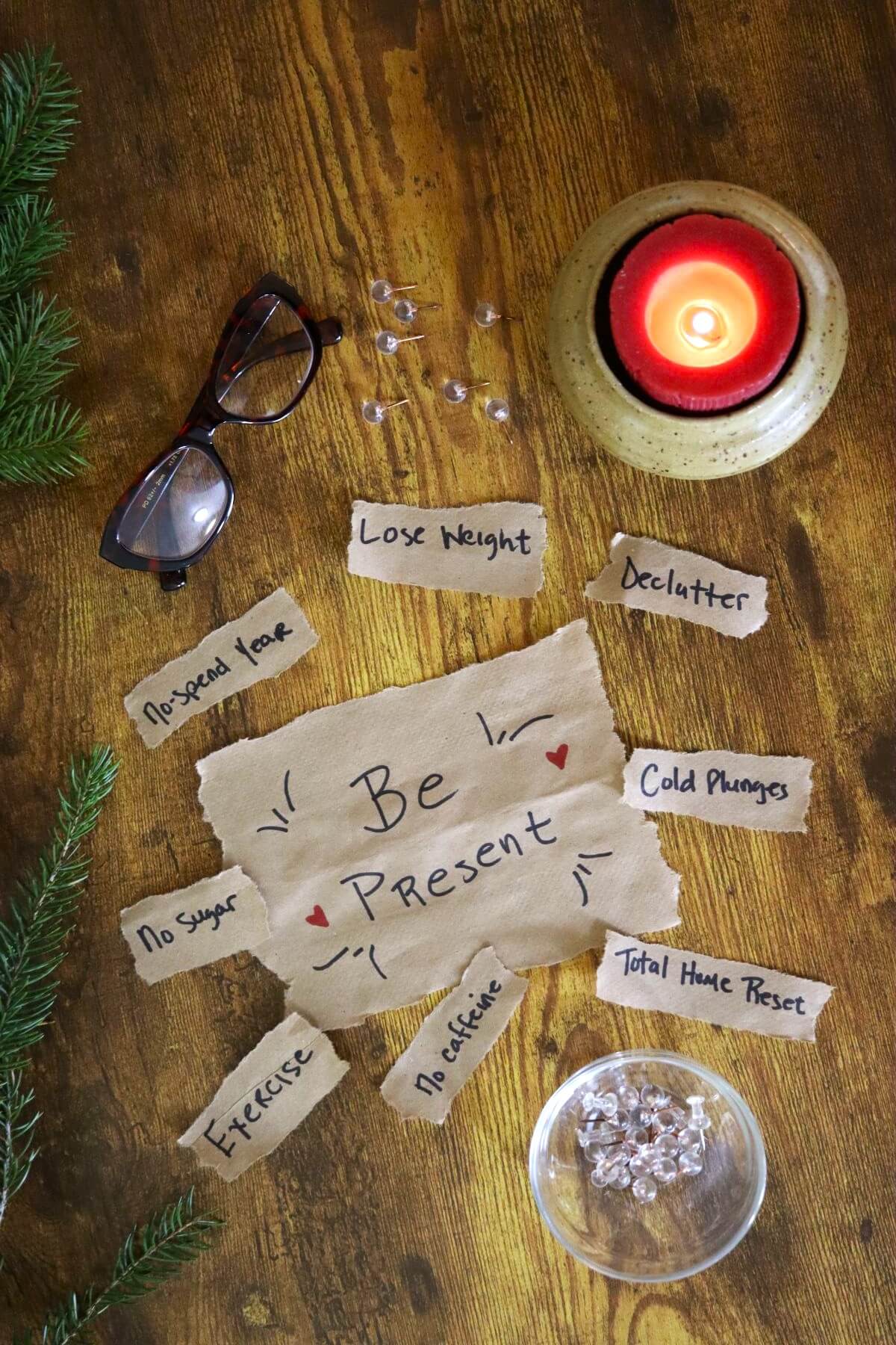 A wooden table with a lit red candle, eyeglasses, greenery, and torn paper slips listing goals like “Lose Weight,” “Declutter,” and “Exercise.” The center paper reads “Be Present” with small hearts drawn beside it.