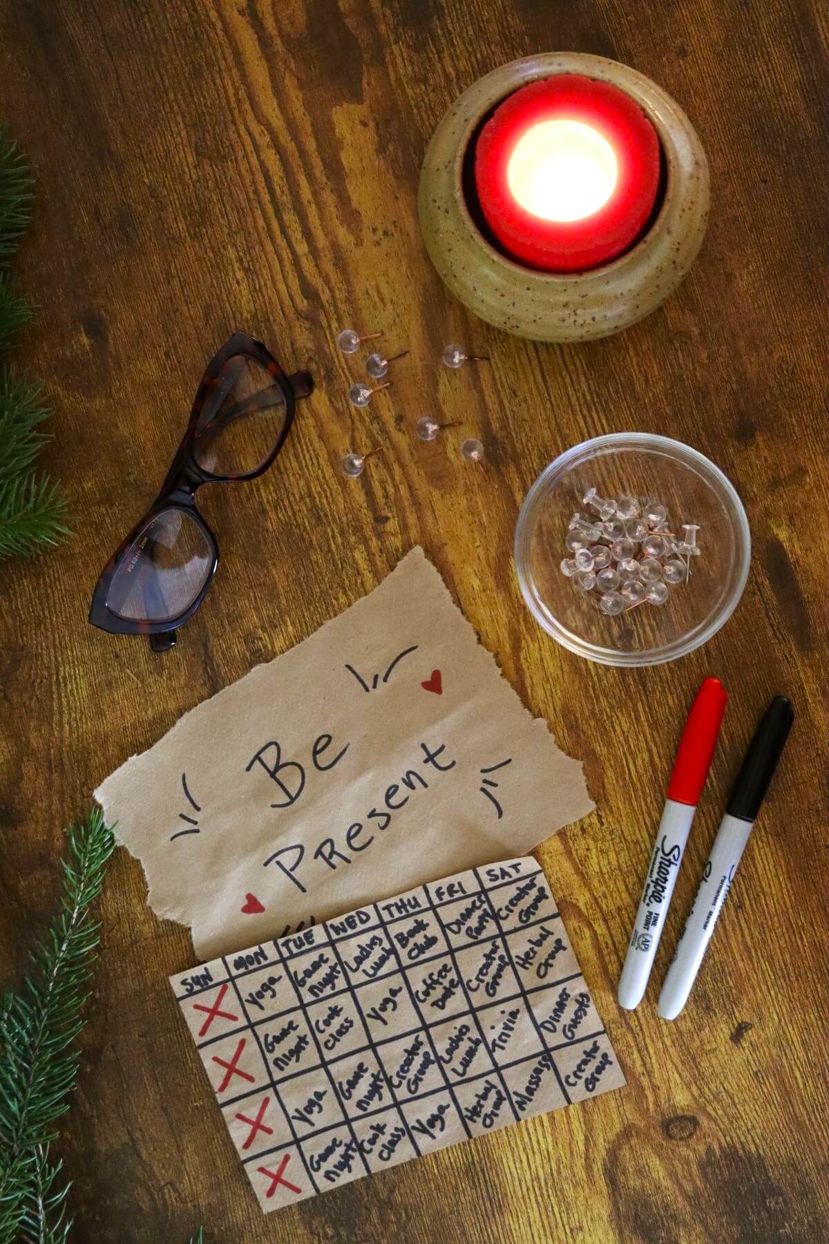 A tabletop with a lit red candle, eyeglasses, two Sharpie markers, a bowl of clear beads, a sprig of greenery, and handwritten notes—one says Be Present, the other is a calendar with tasks and red X marks.