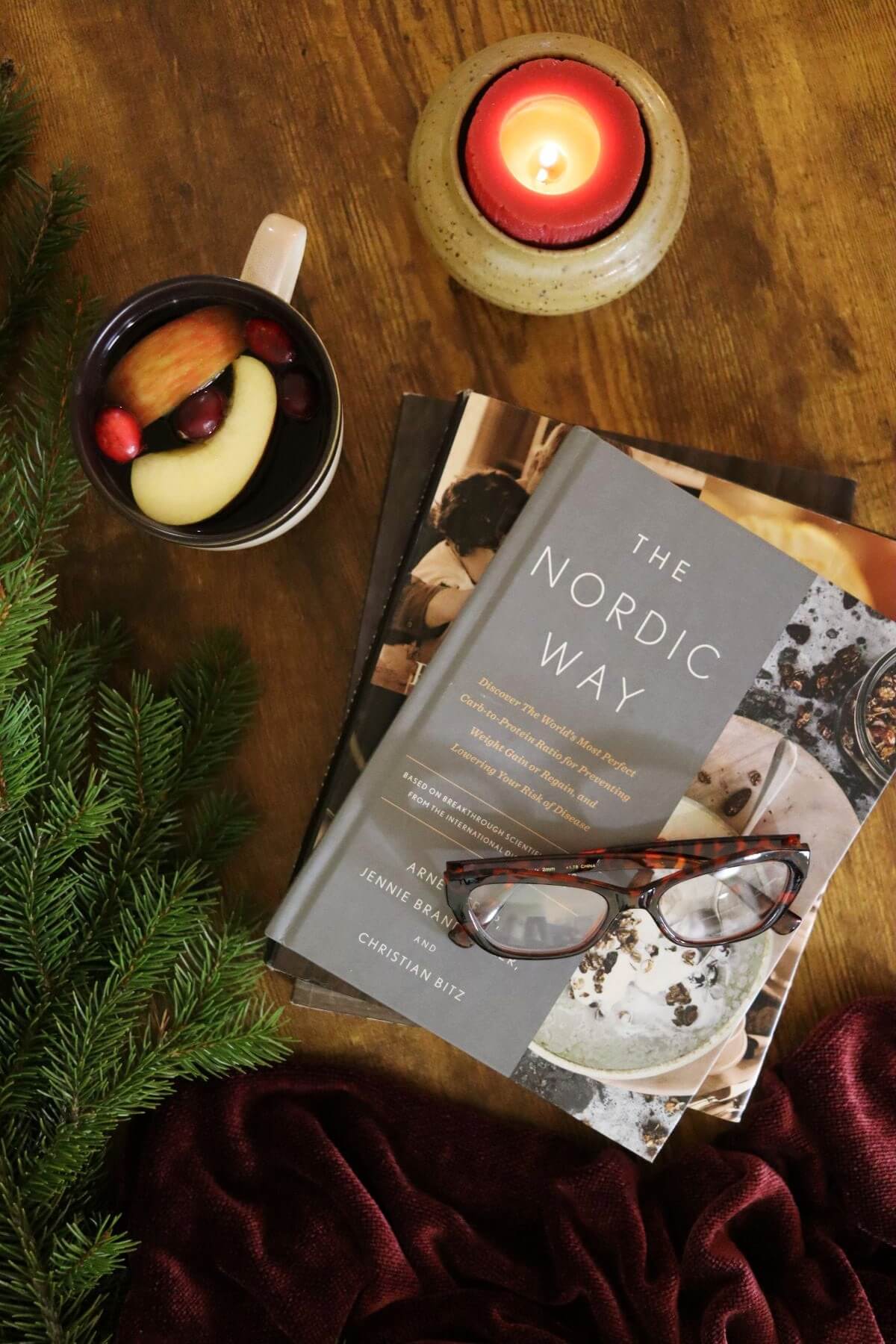 A cozy scene with a candle, a mug of mulled drink with fruit slices, fir branches, a red scarf, and two stacked books — the top one titled The Nordic Way with reading glasses resting on it.