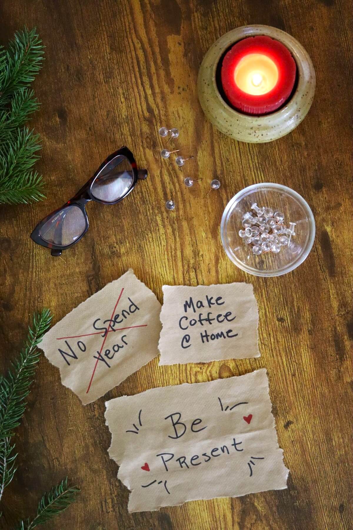 A tabletop with a candle, eyeglasses, fake crystal thumbtacks, evergreen sprigs, and three notes reading: No Spend Year, Make Coffee @ Home, and Be Present, written on torn brown paper.