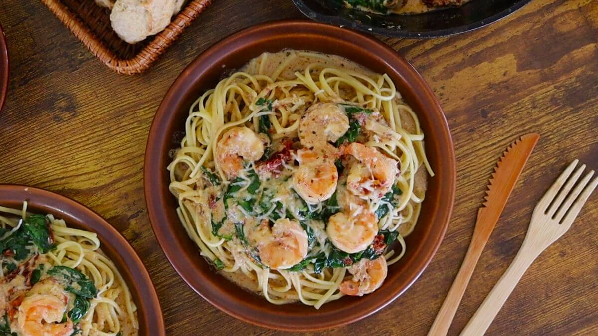 A plate of creamy shrimp pasta with spinach and grated cheese sits on a wooden table, with a basket of bread and a bowl of food in the background. A wooden fork is placed beside the plate.