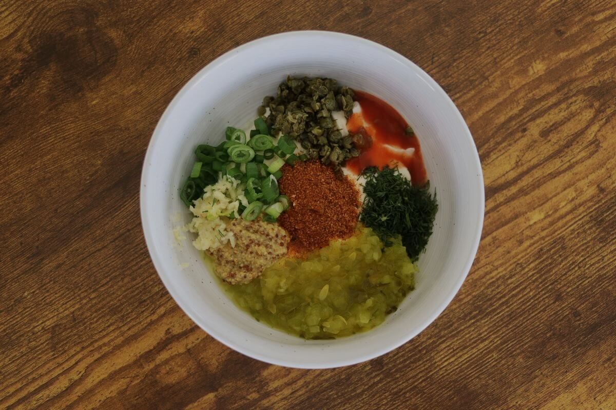 A bowl with mayonnaise, capers, hot sauce, chopped dill, dill relish, mustard, garlic, scallions, and cajun seasoning in it on a wooden surface.