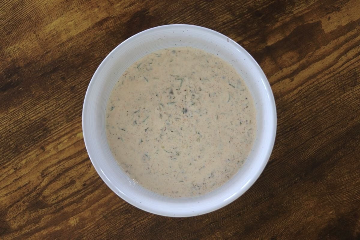 A creamy lightly orange sauce in a white bowl with seasonings visible in it, sitting on a wooden surface.