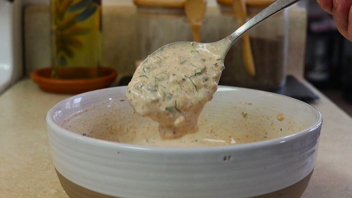 A spoon lifting creamy, herb-filled dip or sauce from a white bowl on a kitchen counter, with containers and a bottle in the blurred background.