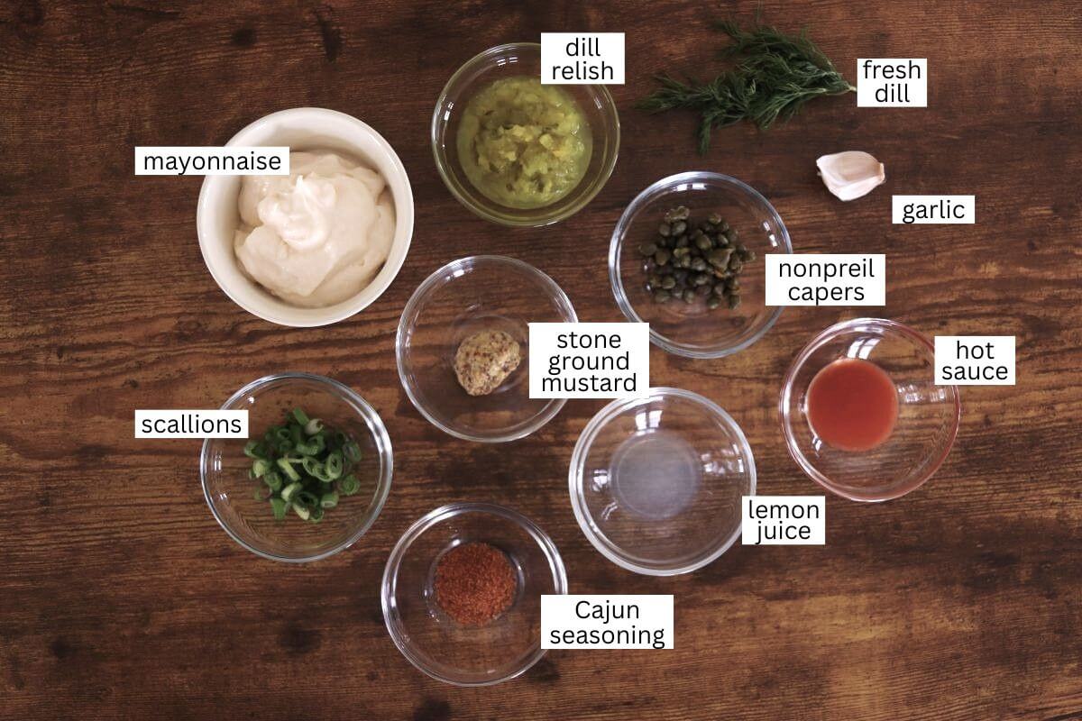 A wooden surface with labeled bowls of mayonnaise, dill relish, fresh dill, garlic, nonpareil capers, hot sauce, lemon juice, stone ground mustard, Cajun seasoning, and scallions arranged neatly.