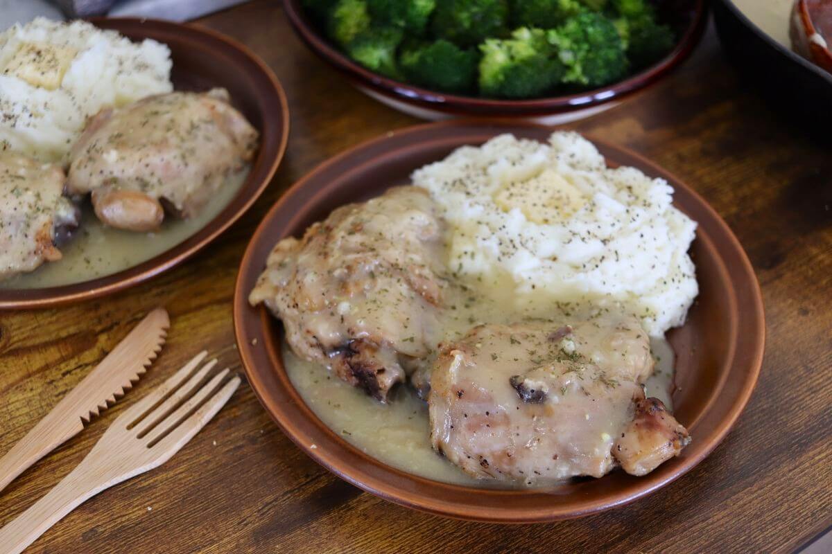 Two chicken thighs covered in gravy on a plate next to mashed potatoes topped with butter and pepper. There is another dish of the same, a dish of broccolit, and a fork and knife visible around the plate.