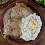 Two chicken thighs smothered in gravy sit on a plate next to mashed potatoes topped with a chunk of butter and sprinkled with pepper.