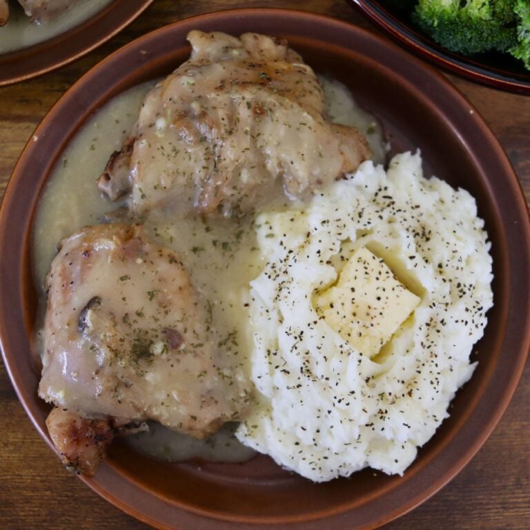 Two chicken thighs smothered in gravy sit on a plate next to mashed potatoes topped with a chunk of butter and sprinkled with pepper.