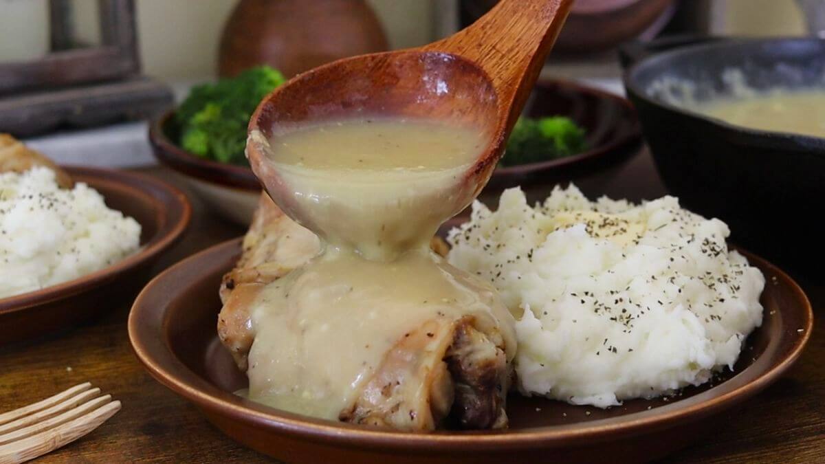 A wooden spoon pours creamy gravy over a piece of cooked chicken, served next to a mound of mashed potatoes with black pepper on a brown plate. Broccoli and sauce are visible in the background.