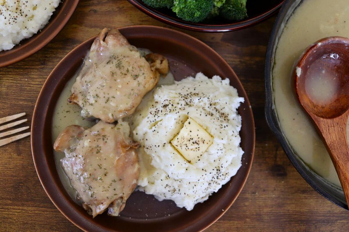 A plate with two pieces of baked chicken covered in gravy, served next to mashed potatoes topped with a pat of butter and sprinkled with black pepper. A fork, skillet of gravy, and bowl of broccoli are nearby.