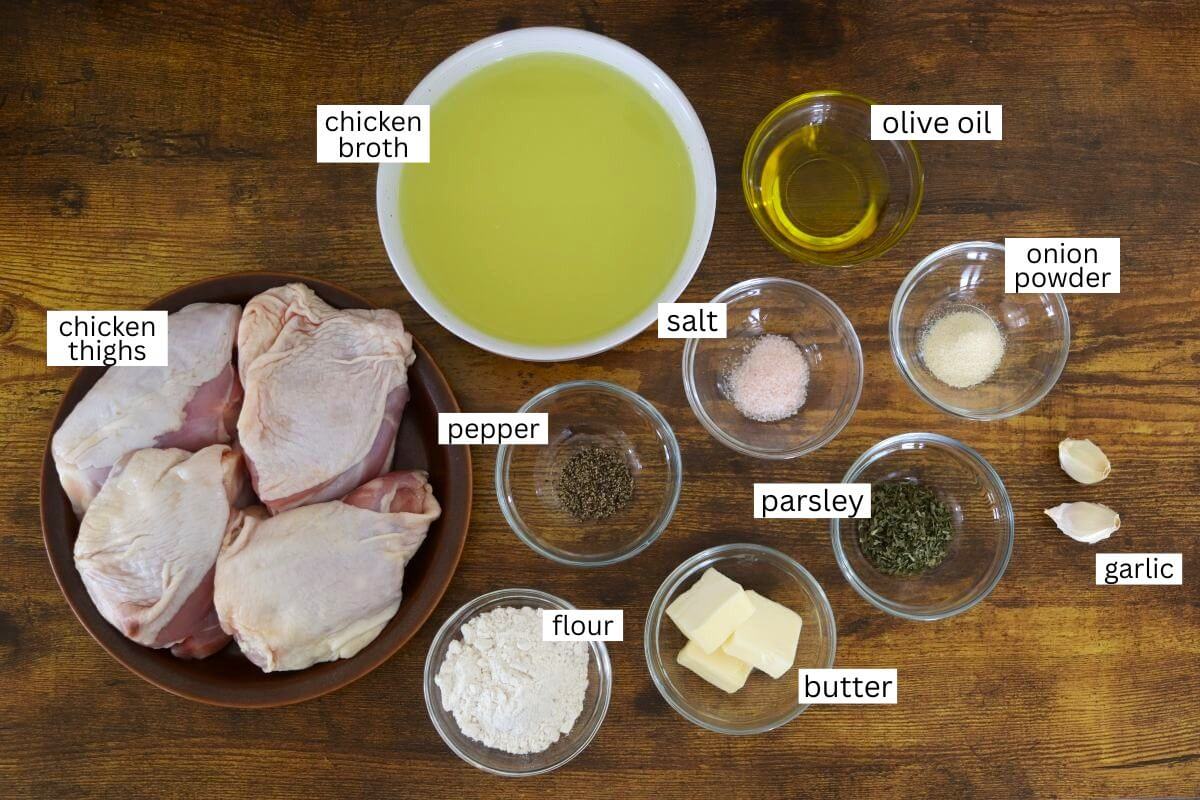 Ingredients on a wooden surface: raw chicken thighs, chicken broth, olive oil, salt, pepper, onion powder, dried parsley, garlic cloves, butter, and flour, each labeled in a small bowl or dish.